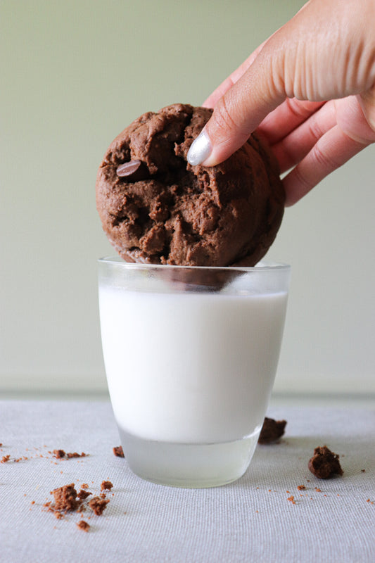 Chocolate cookie being dunked into a glass of milk with a hand holding the cookie.